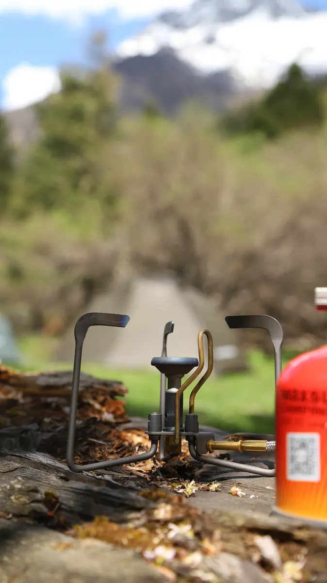 Close-up of the Petrel Summit burner on a fallen log with its pot supports open, a gas canister beside it and a mountain ridge out of focus behind.